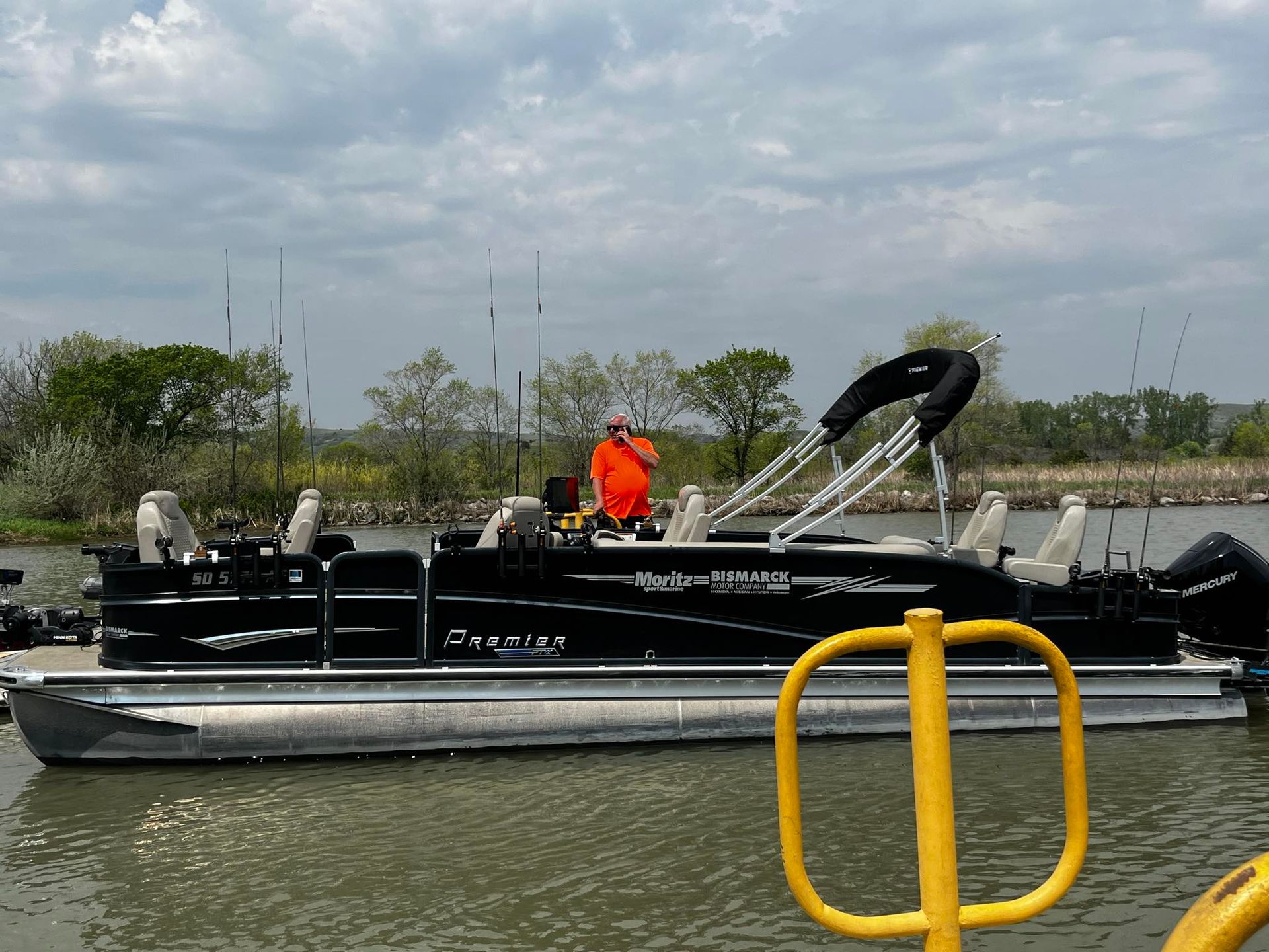 Black pontoon boat with a person at the helm on the water; overcast sky.