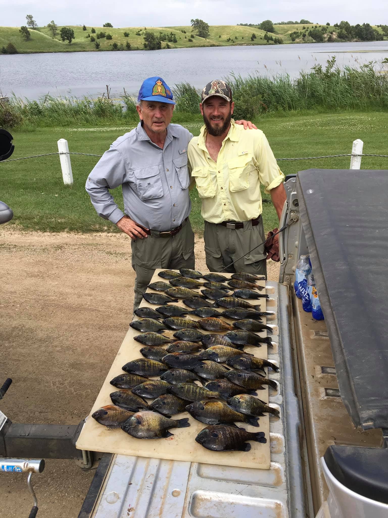 Two men pose with a large catch of fish on a tailgate near a lake.