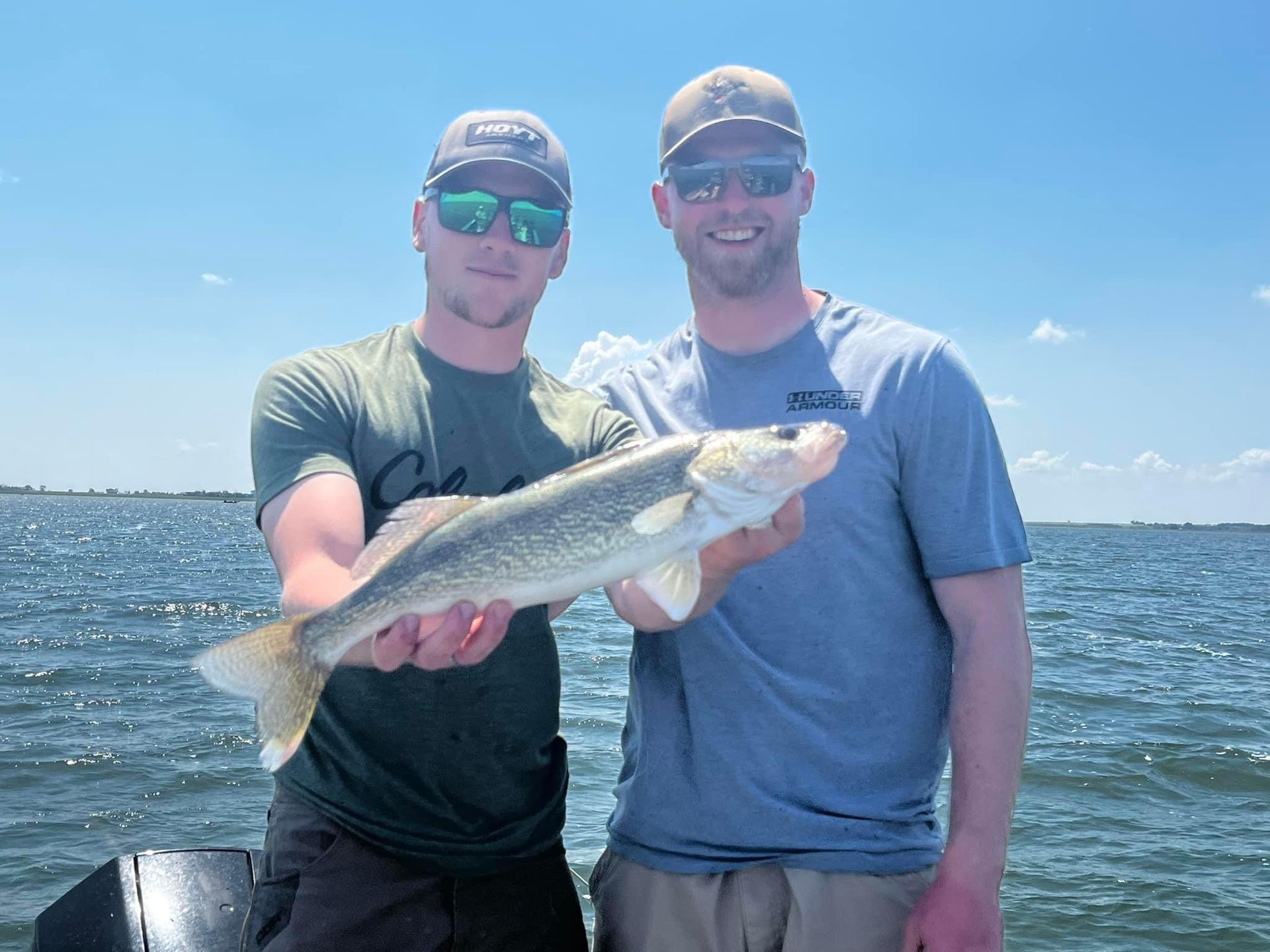 Two men on a boat hold up a fish, posing for a photo. Blue sky and water are in the background.