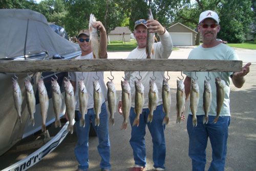 Three people display caught fish, hanging on a wooden rack next to a boat on a sunny day.