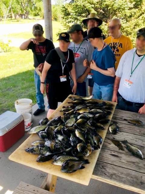 Group of people gathered around a table piled with fish, likely after a fishing trip, outdoors.