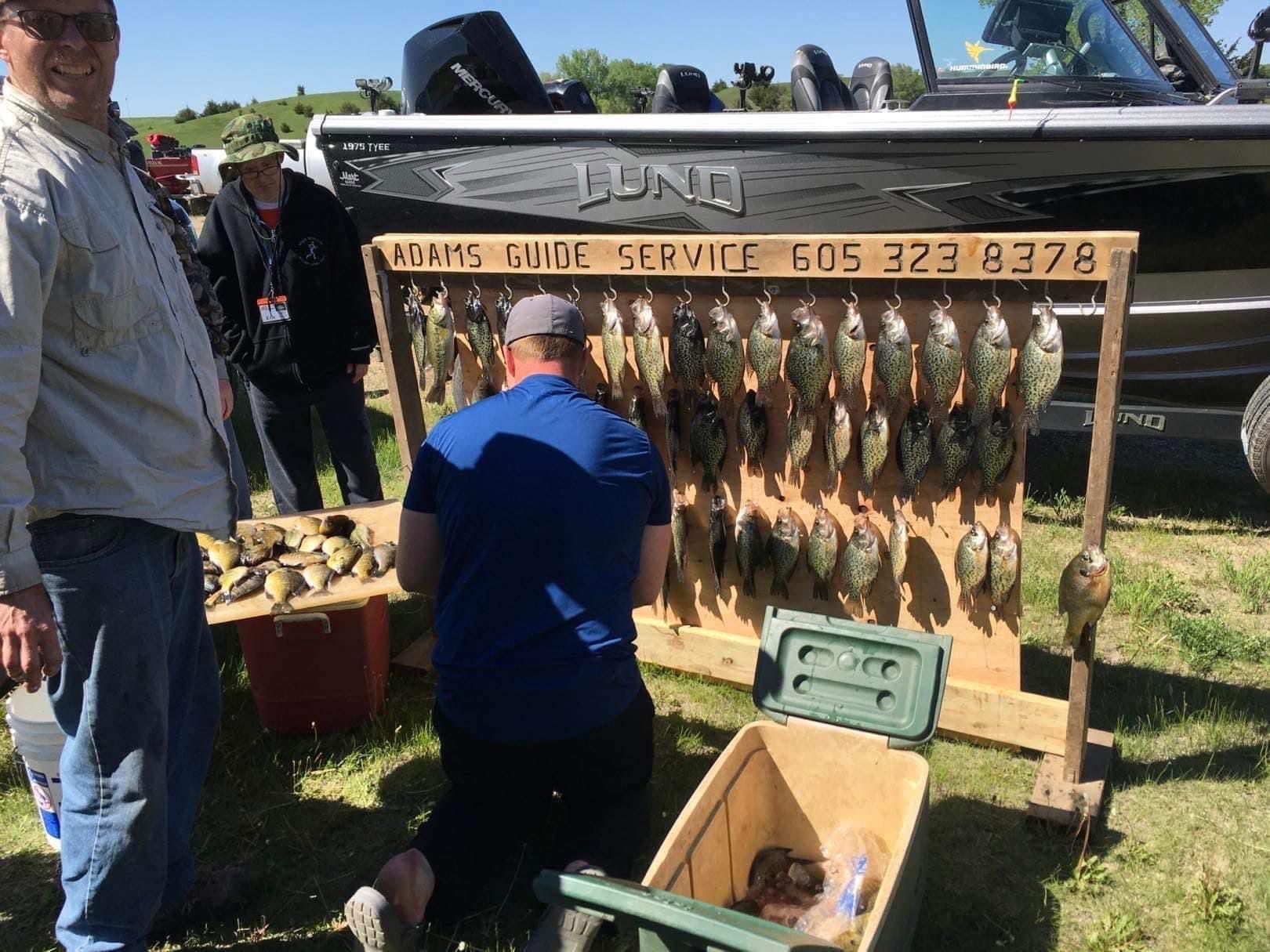 People near a wooden rack displaying numerous fish, Adams Guide Service, a fishing guide service. A boat is in the background.