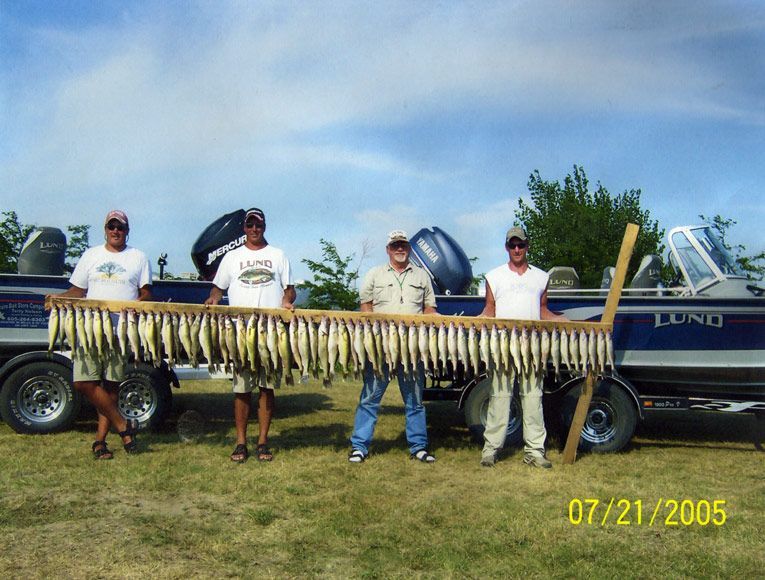 Four men holding a wooden frame displaying a large catch of fish near a boat on a grassy field; cloudy sky.
