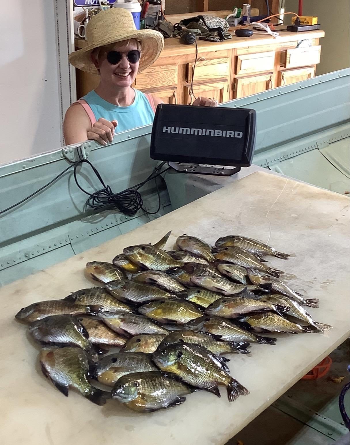 Woman smiling, holding a fish finder, and showing off a pile of caught fish in a boat.