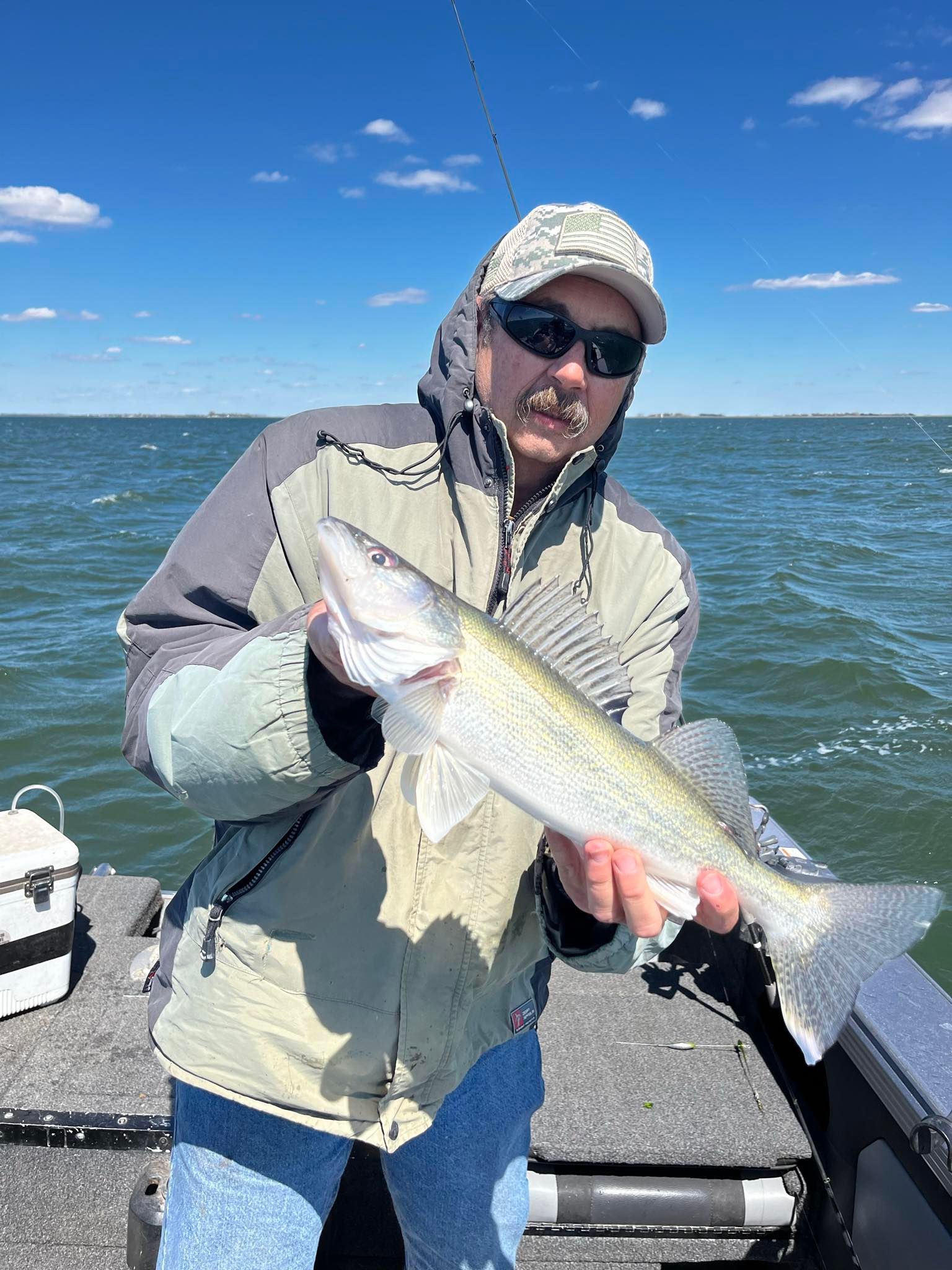 Angler holding a walleye on a boat, wearing sunglasses and jacket; blue sky, water.