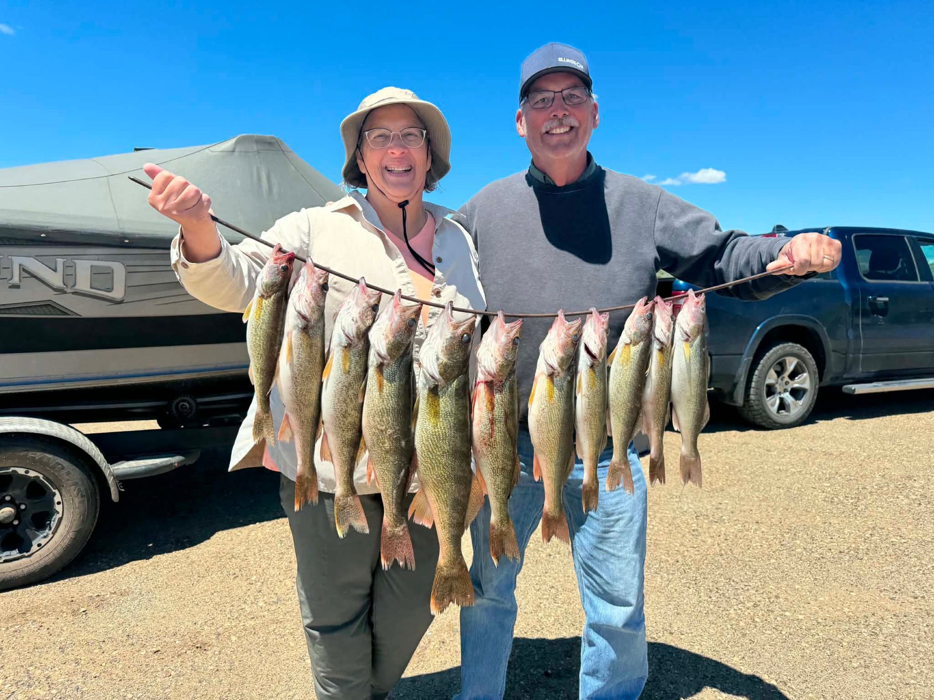 Two people smiling and holding a string of fish beside a truck under a blue sky