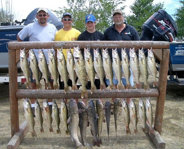 Four men stand behind a wooden rack holding many freshly caught fish near a lake.