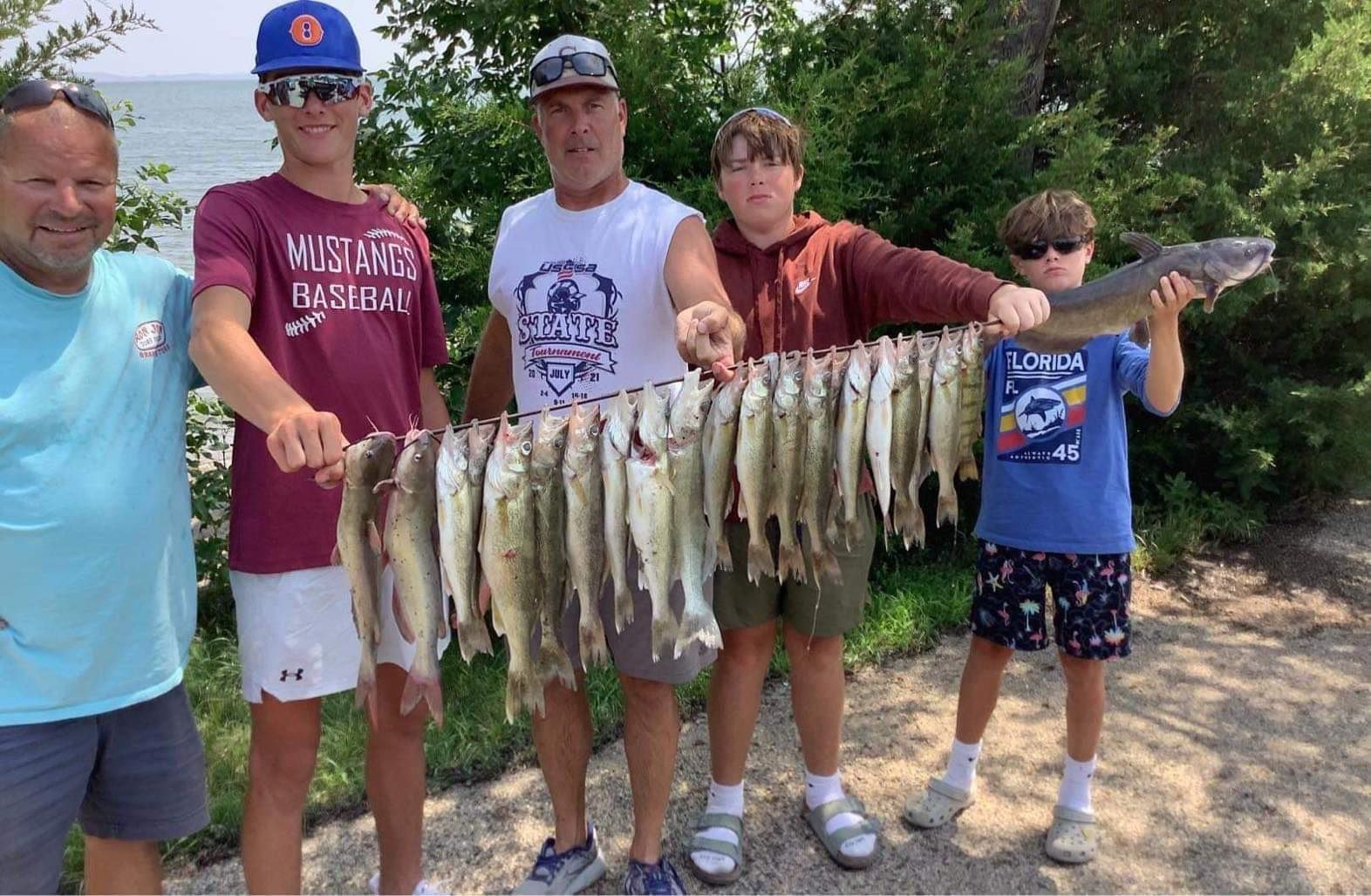 Five people holding up a string of caught fish near a body of water. Sunny day.