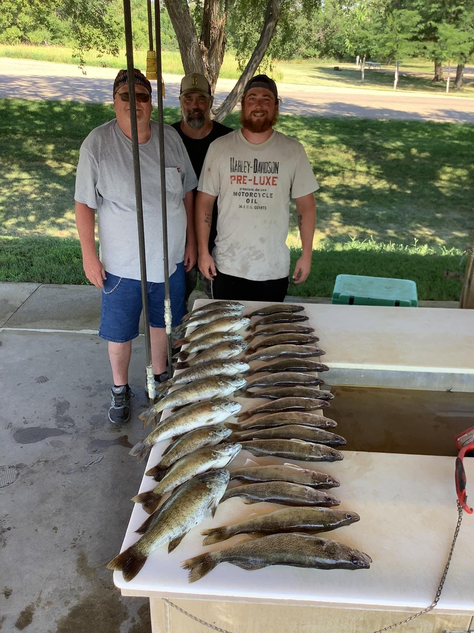 Three people stand behind a table displaying a large catch of fish; outdoor setting.