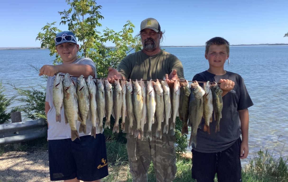 Three people holding up a string of caught fish near a body of water on a sunny day.
