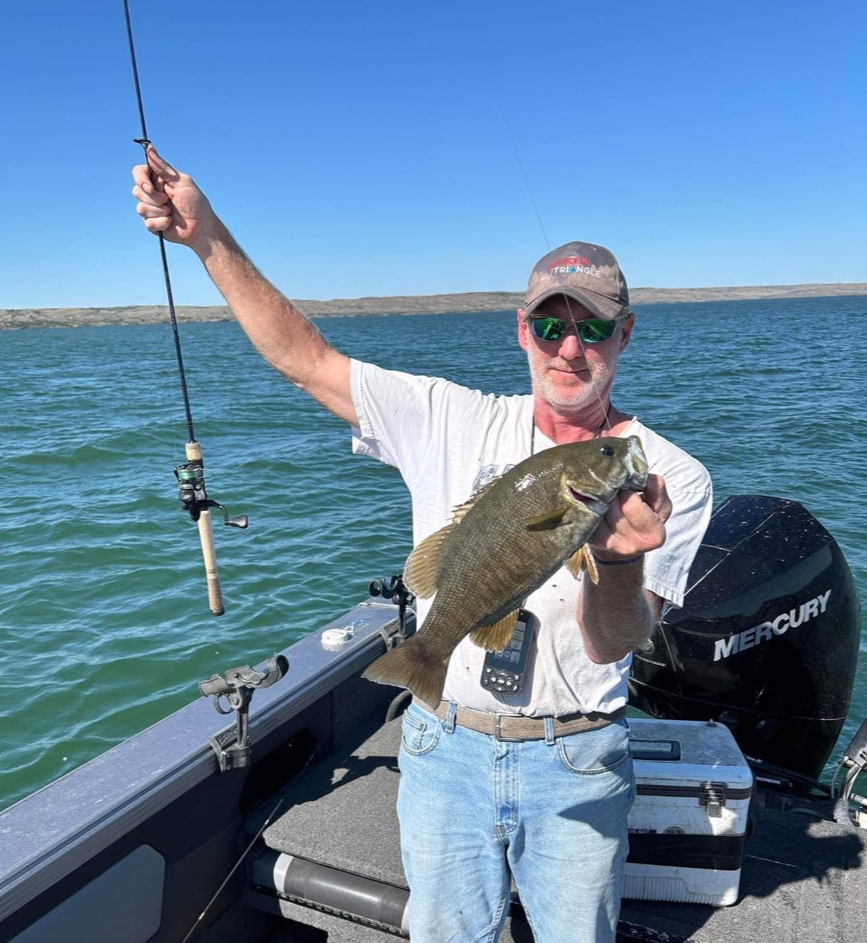 Man on a boat holds up a fish, fishing rod in the air, against a blue sky.