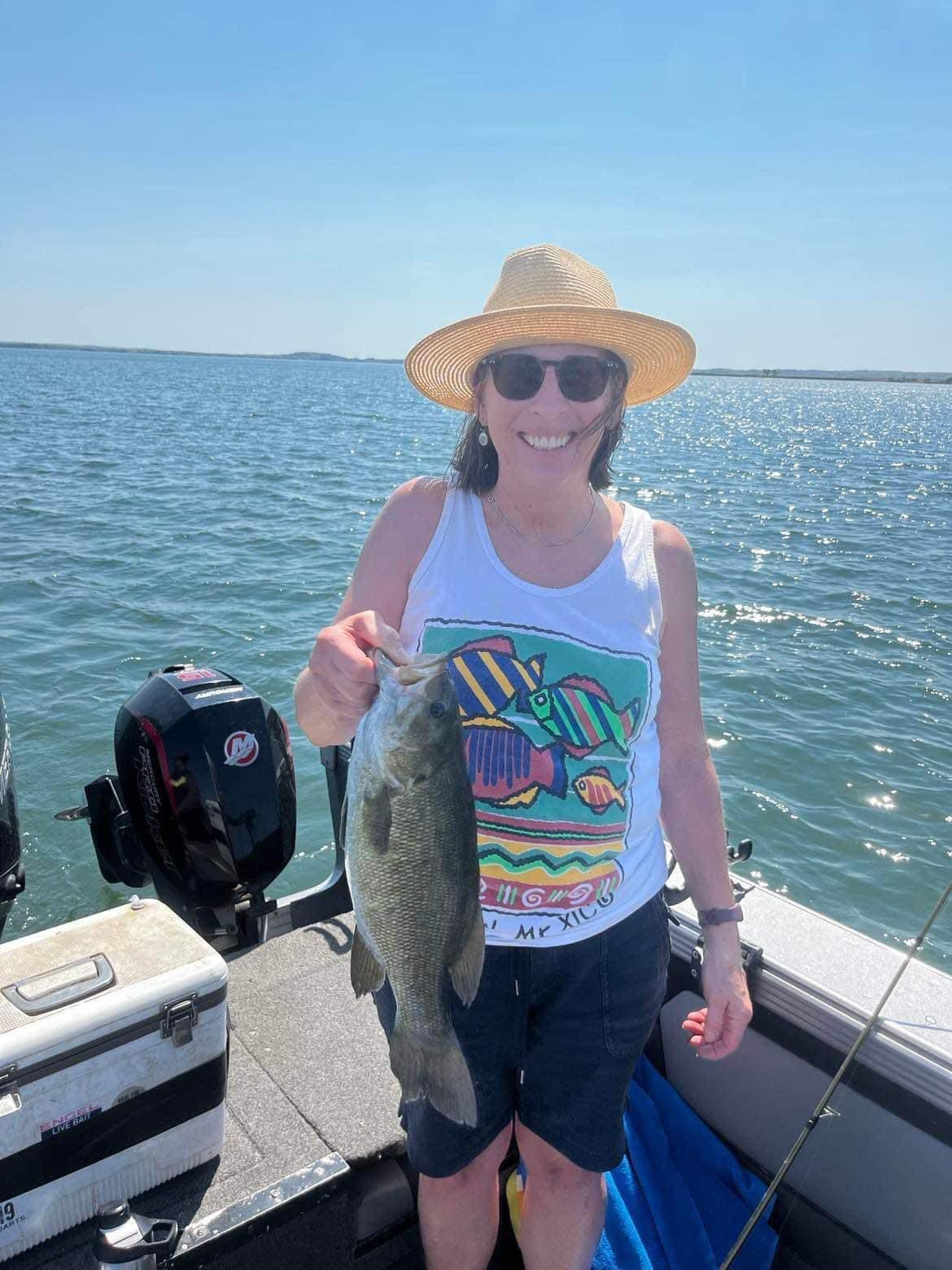 Woman on boat holding fish, wearing hat and sunglasses, sunny day.