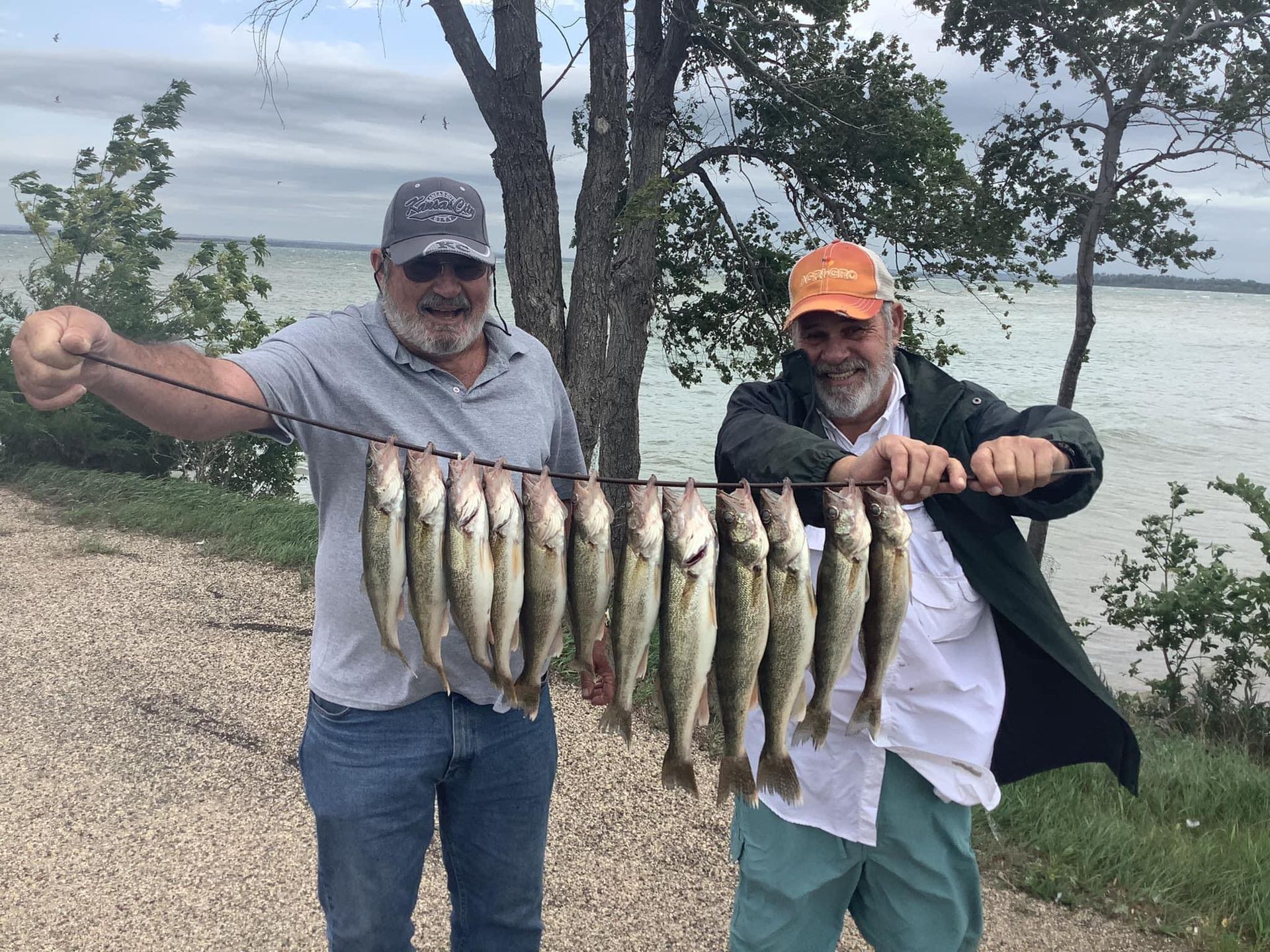 Two men holding a string of fish they caught by a body of water. Overcast day.