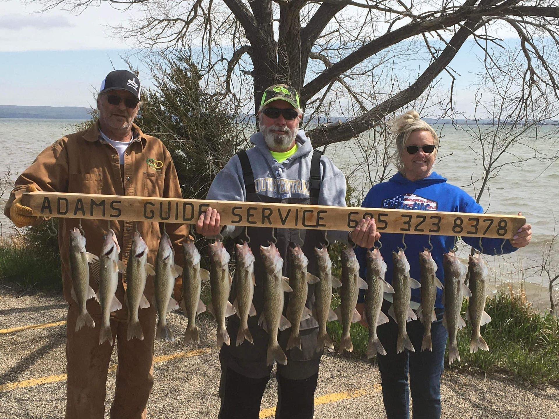 Three people holding a wooden sign and a string of caught fish near a body of water.