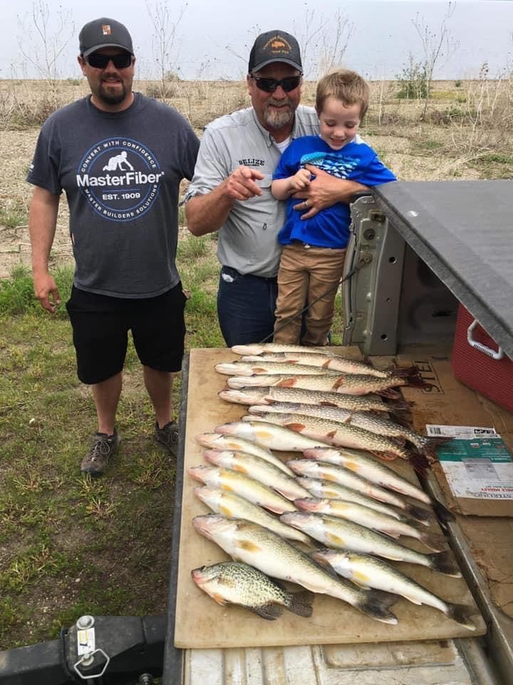Three people beside a truck bed display a large catch of fish on a cutting board. The man in the middle points at the fish.
