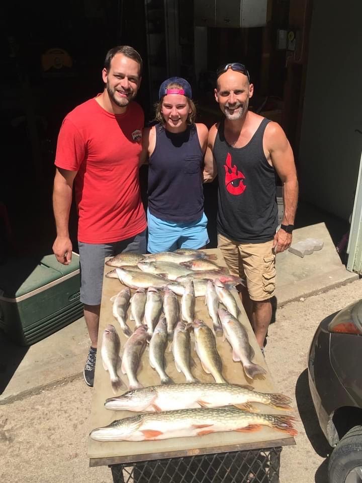 Three people pose with a large fish catch on a table outdoors; sunny day.