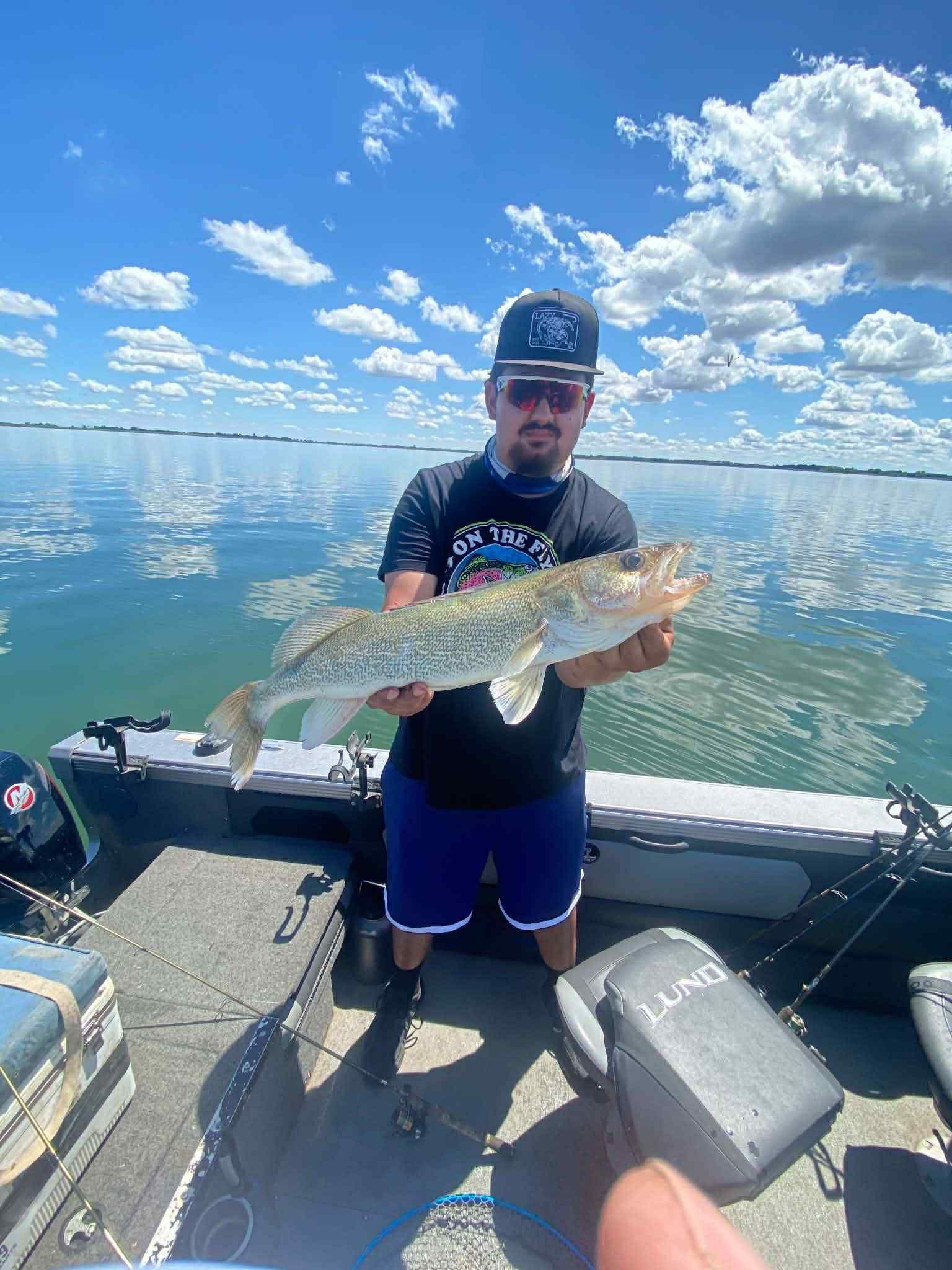 Angler on a boat holds a large fish against a bright blue lake and cloudy sky
