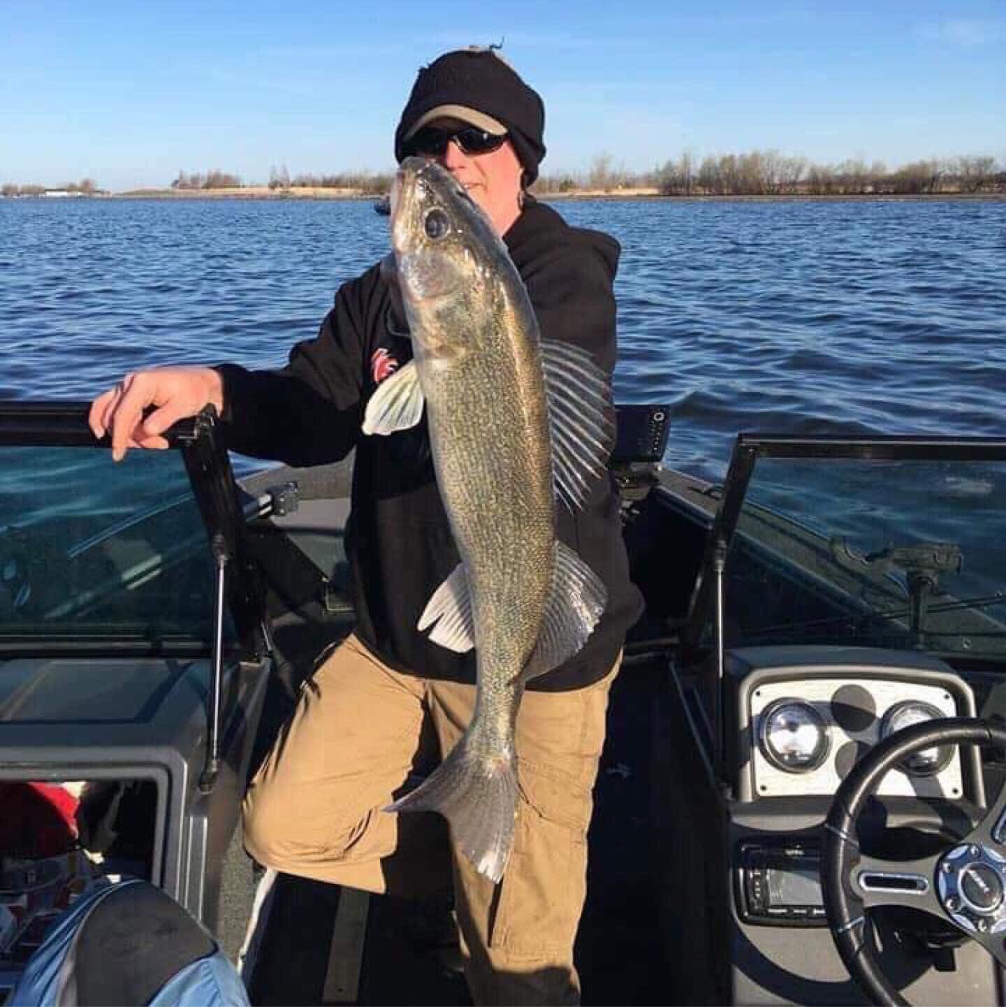 Man holding large fish on a boat; water and shoreline in background.