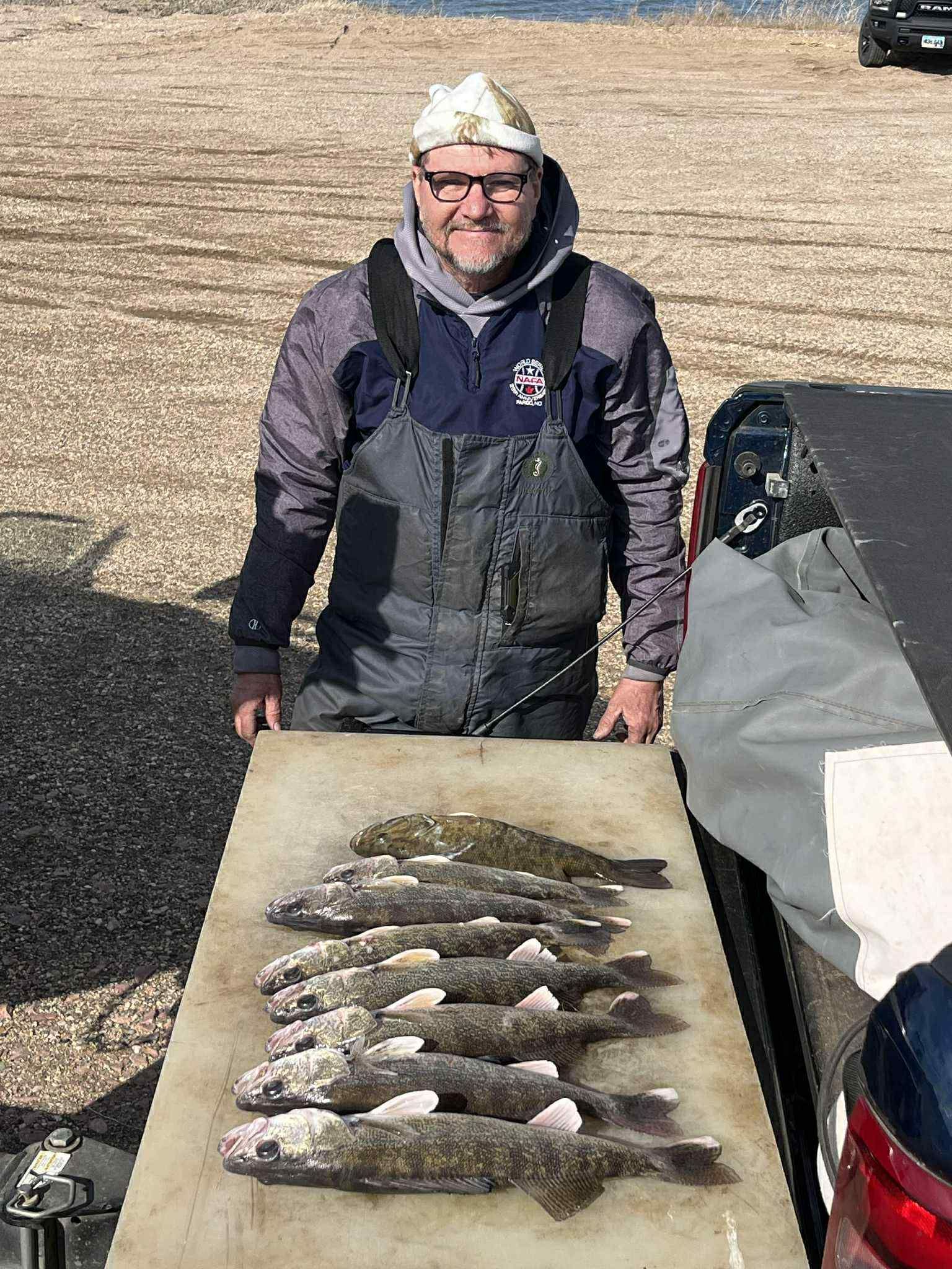 Man in fishing gear displaying a catch of fish on a table near a body of water.