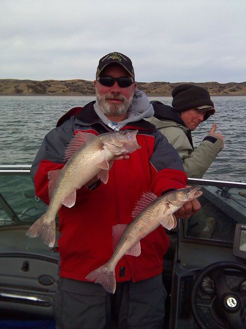 Man holding two fish on a boat; another man gives a thumbs up; water and shore in the background.