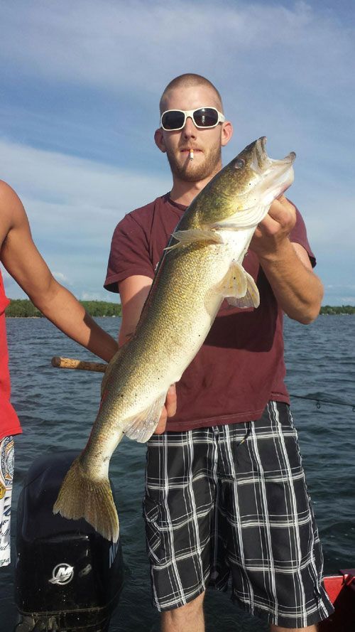 Man holding a large fish on a boat, smoking. Background is a lake and sky.