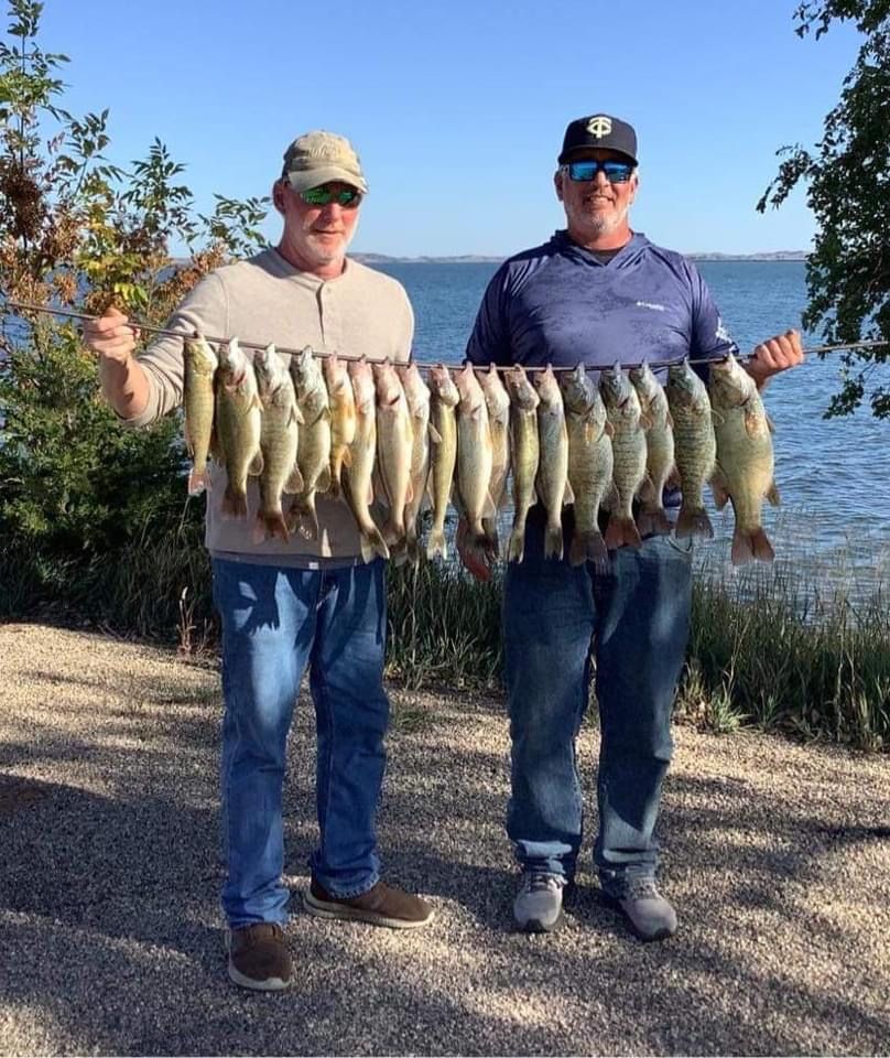 Two men holding up a string of caught fish near a lake.