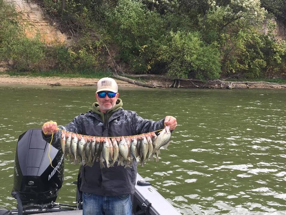 Man holding a stringer of caught fish on a boat in a river with a wooded bank in the background.