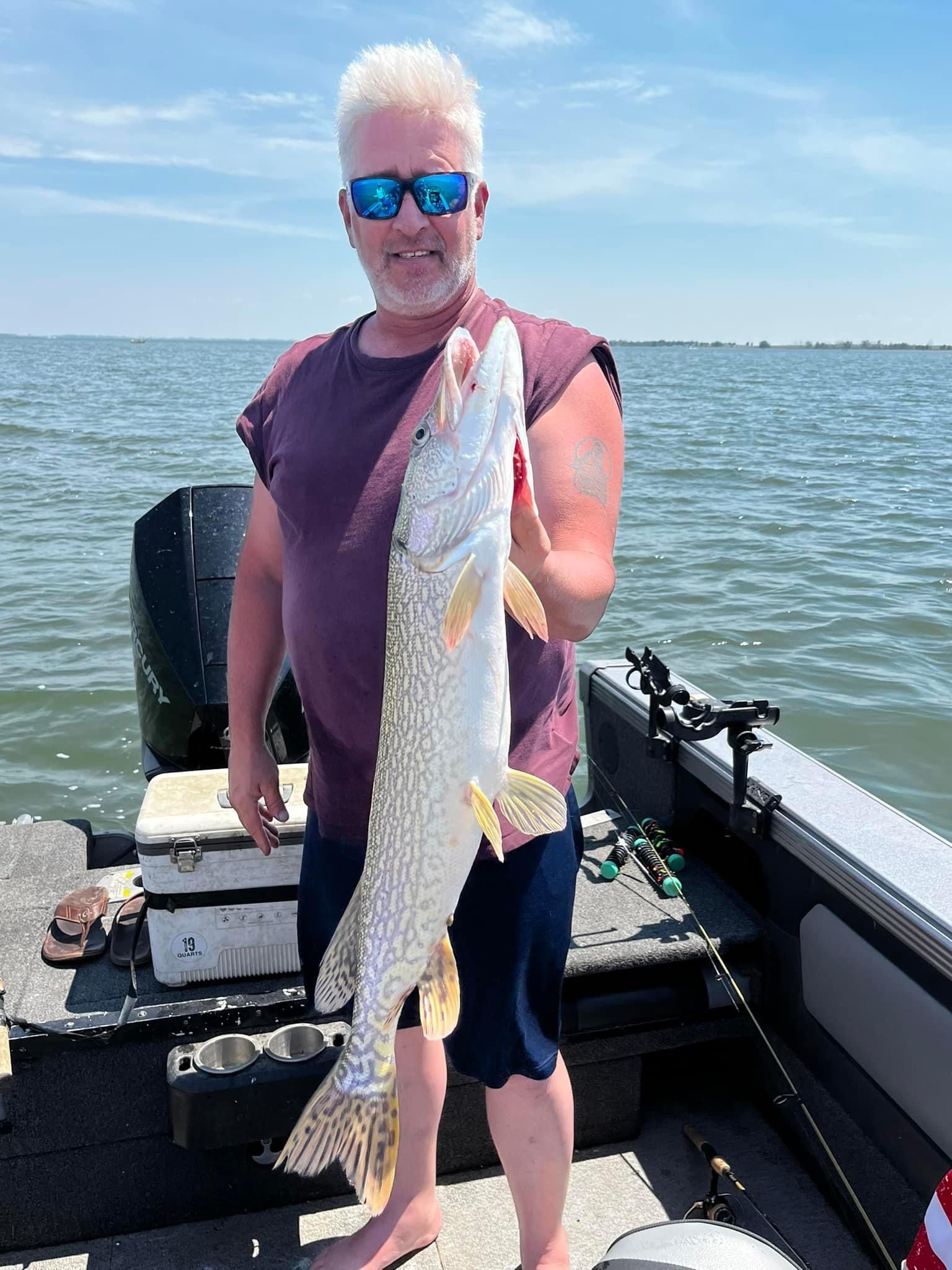 Man holding a large fish on a boat, blue water and sky in background.