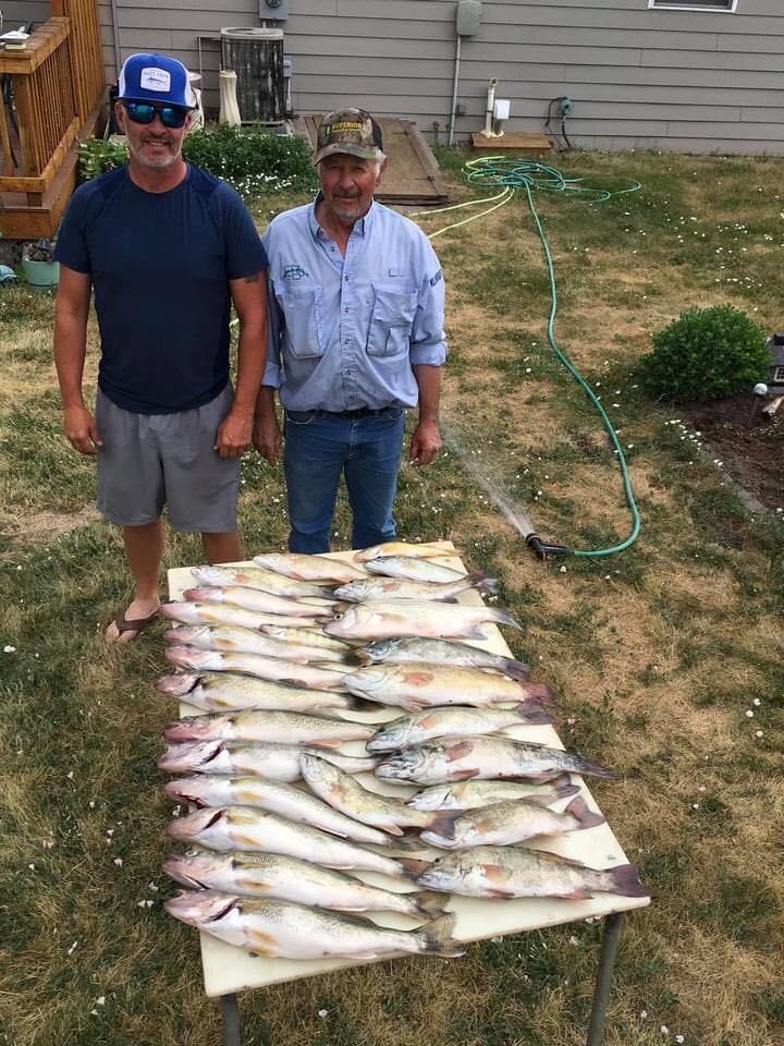 Two men standing behind a table piled with many fish in a backyard setting.