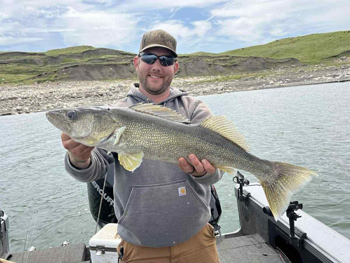 Man on a boat holds up a large walleye fish, smiling. Outdoors, lake and hills in the background.