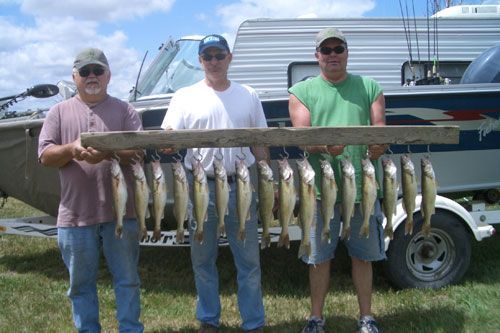 Three men holding a board with many fish hanging from it in front of a boat.