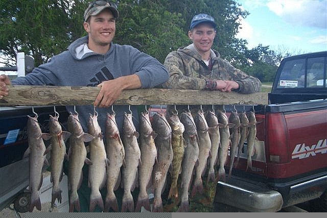 Two men holding a wooden beam with many catfish hanging on it, next to a red pickup truck.