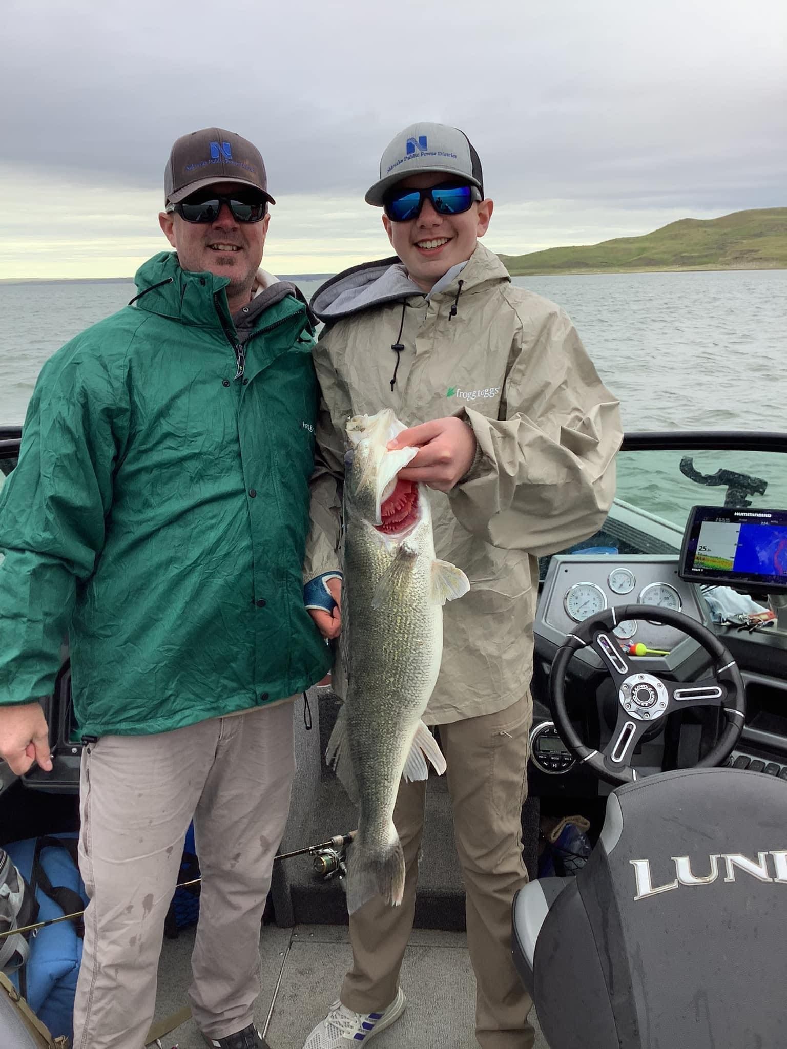 Two people on a boat holding a large fish; person on right is smiling. Overcast sky and water in the background.