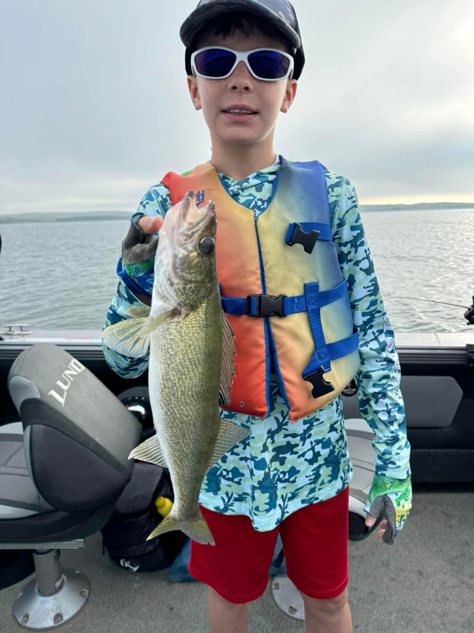 Boy in life vest holding a walleye fish on a boat. Cloudy sky over water.