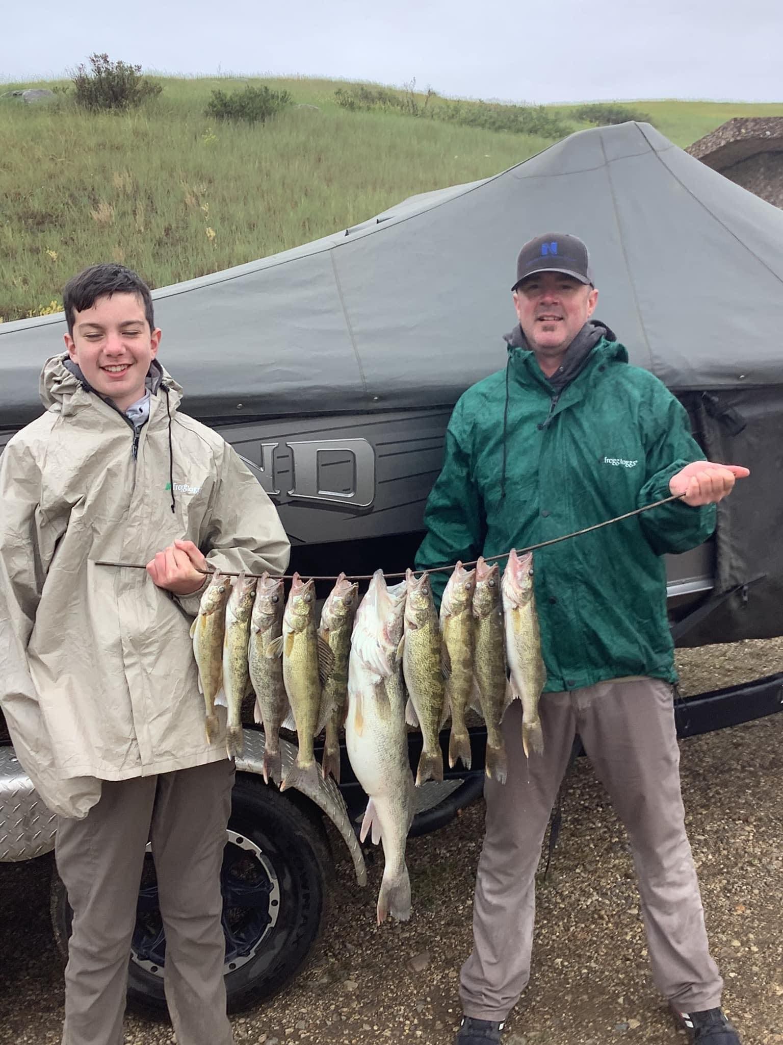 Two people holding a string of caught fish in front of a trailer and grassy hill.
