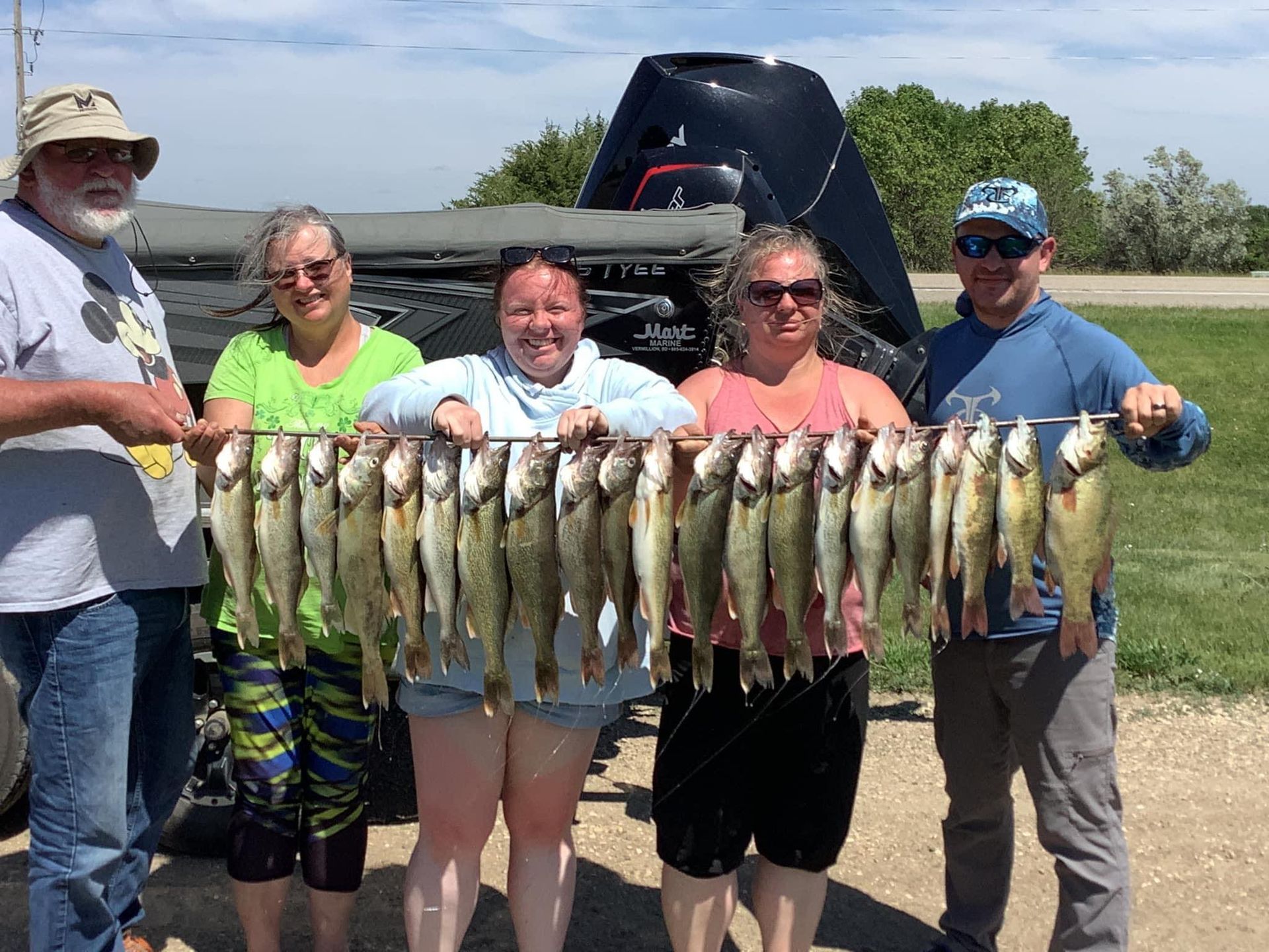 Five people pose with a string of many caught fish in front of a boat. Sunny day.