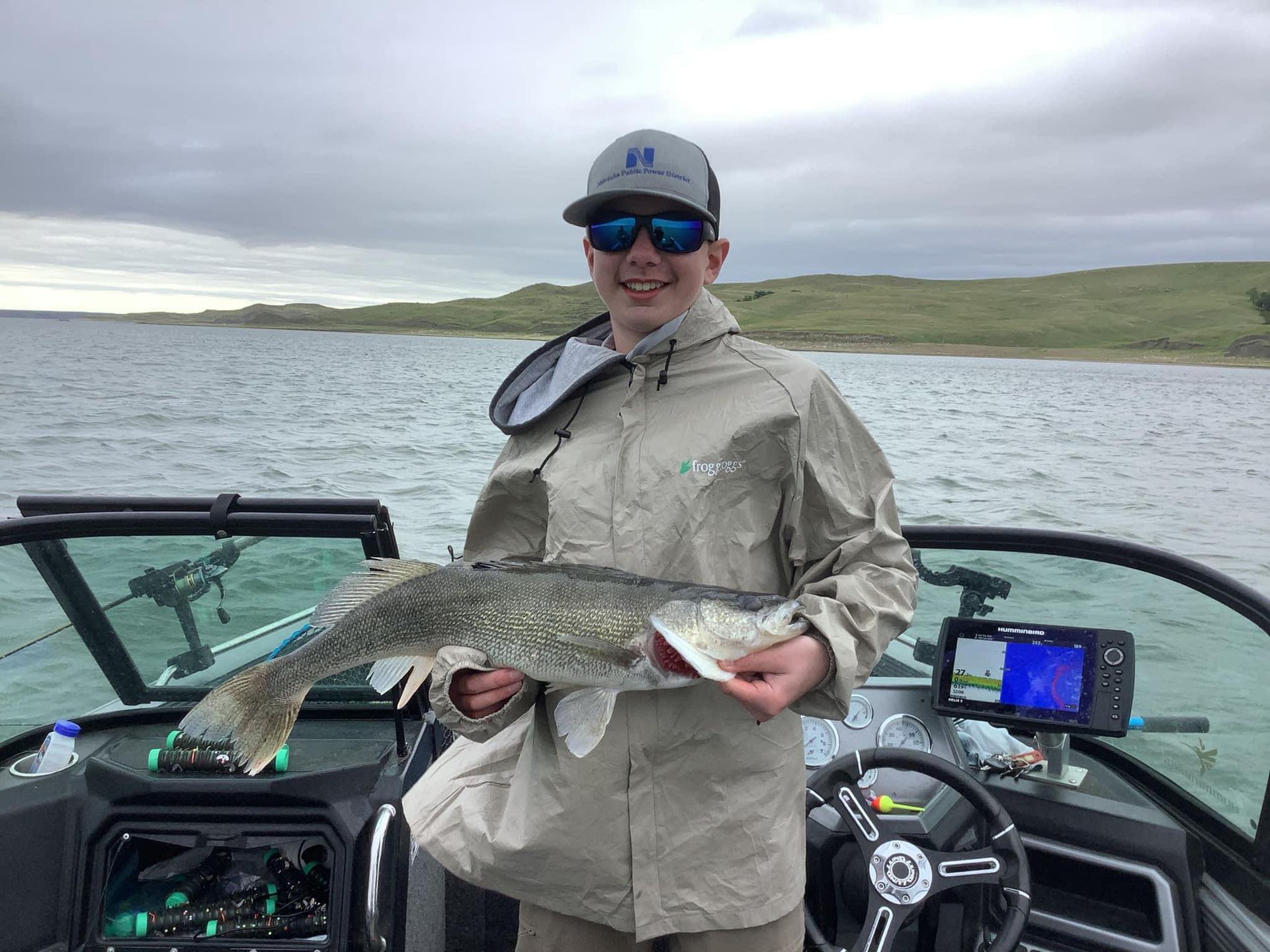 Man on a boat holding a fish, near a lake. He wears sunglasses, a hat, and a jacket.