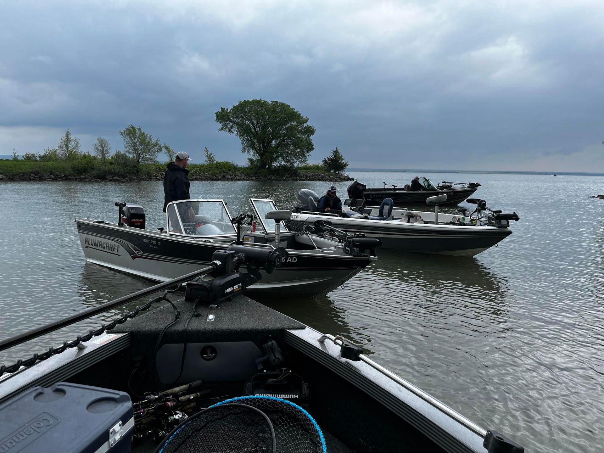 Three fishing boats on water, overcast sky. Anglers in boats, one standing.
