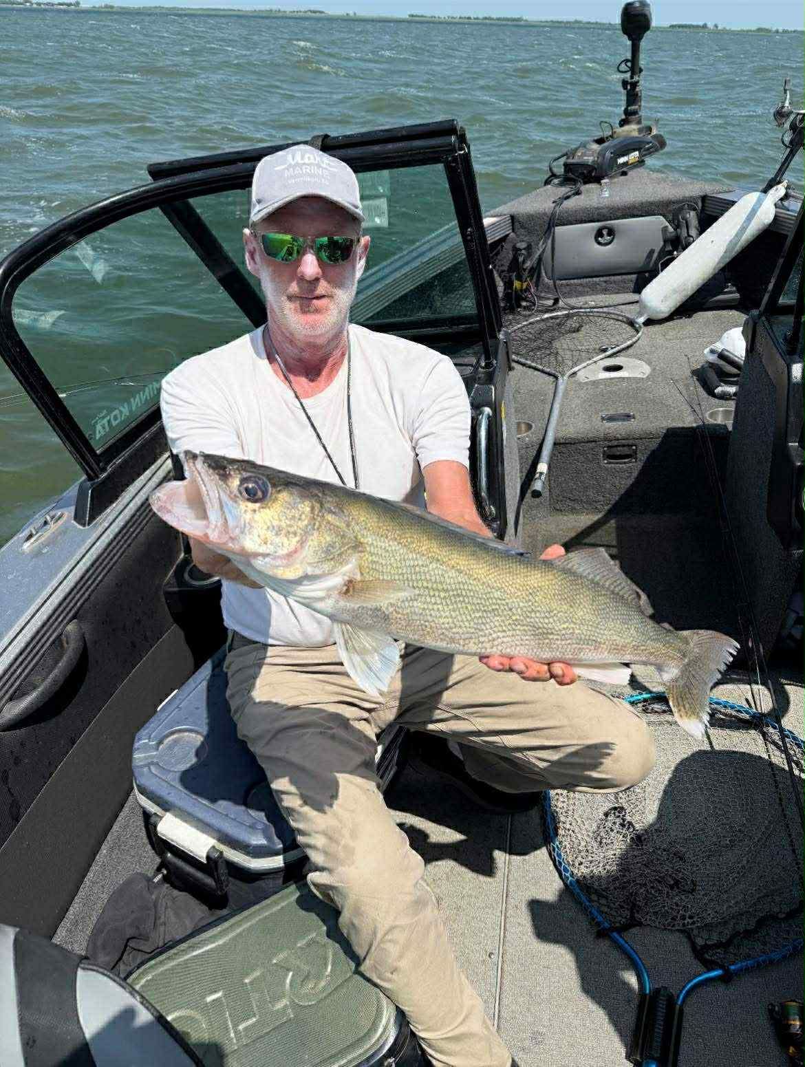 Man holding a large walleye fish on a boat. He's wearing a hat and sunglasses; boat on a lake.