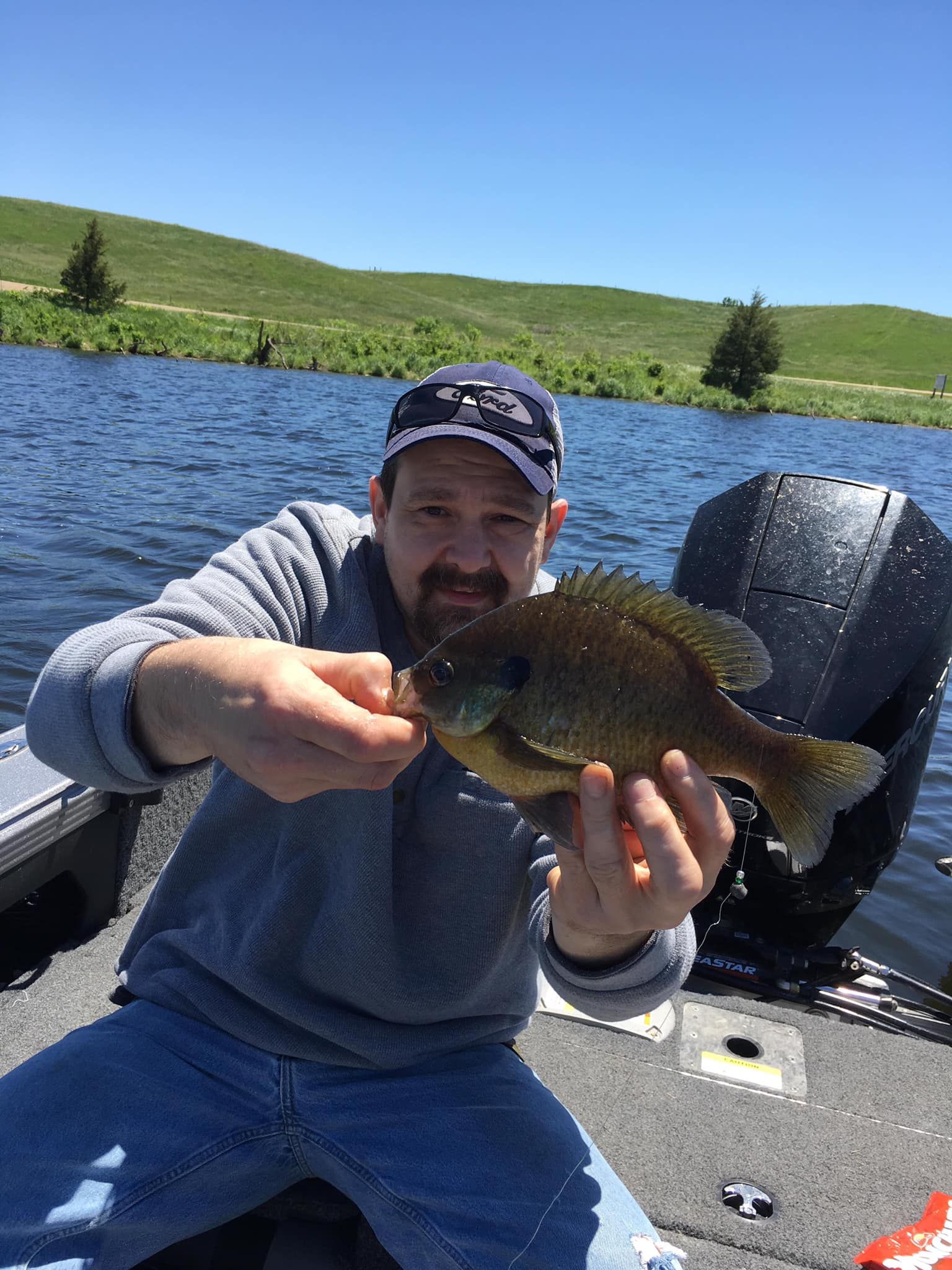 Man on a boat holds up a sunfish, with a lake and hills in the background on a sunny day.