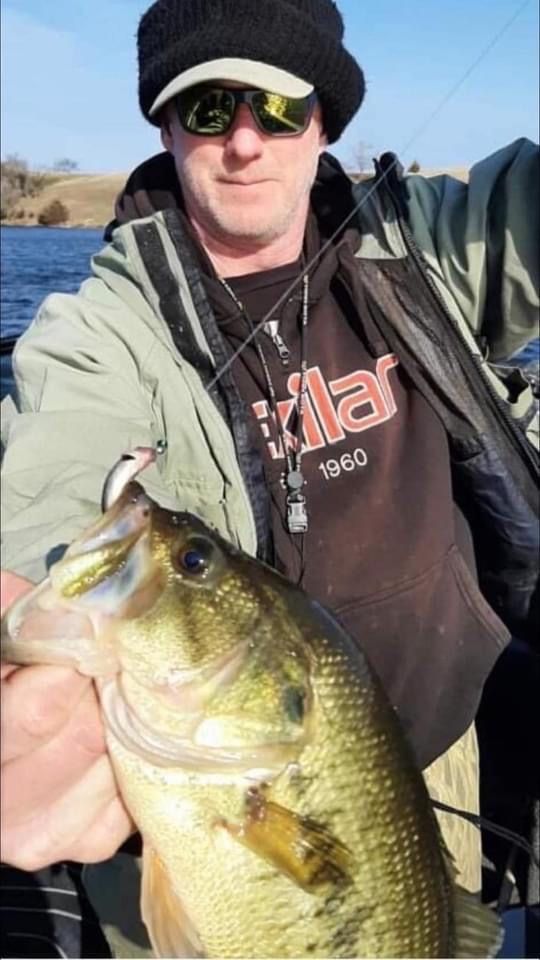 Man holding a large bass, wearing a hat, sunglasses, and a sweatshirt on a boat.