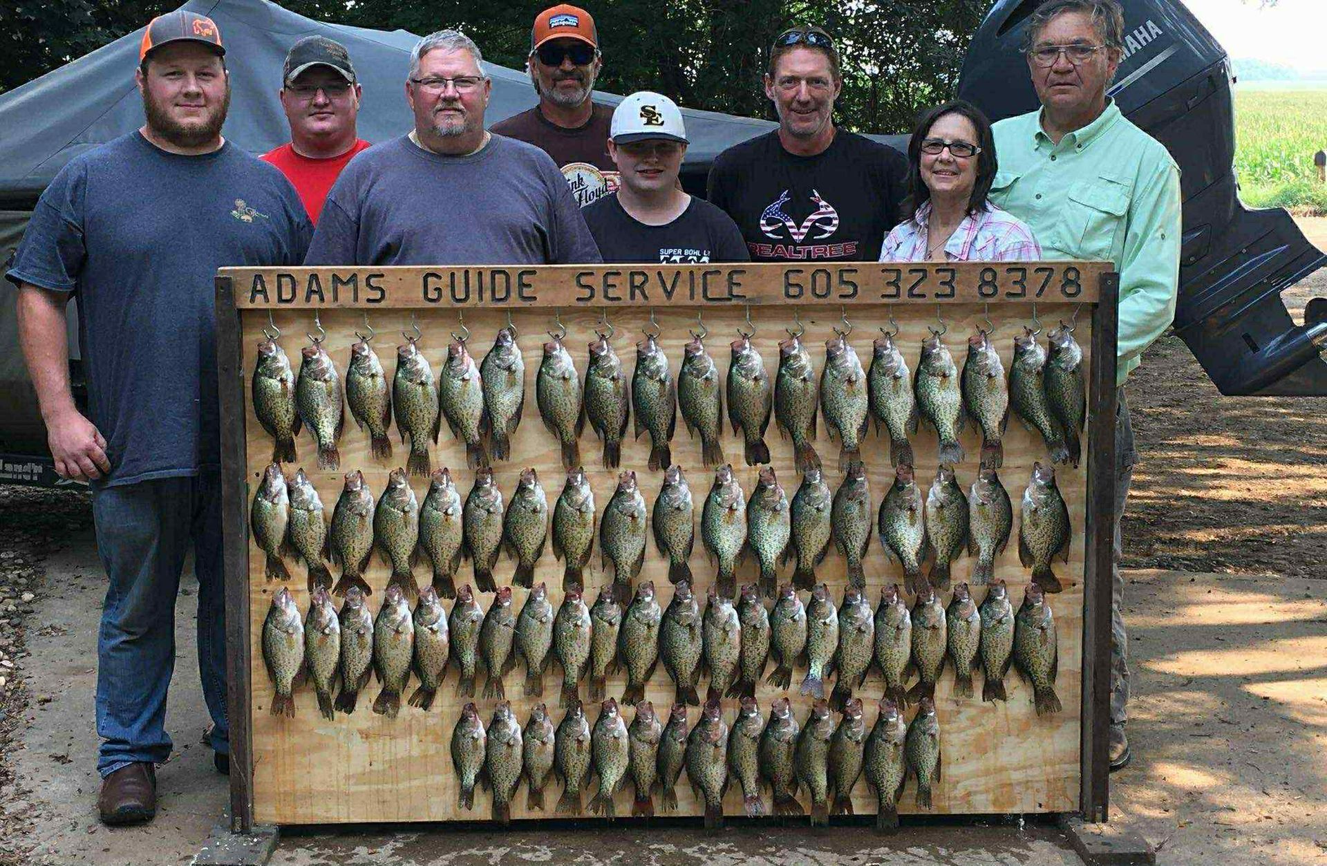 Group of people with a large wooden board displaying many caught fish; outdoor setting.