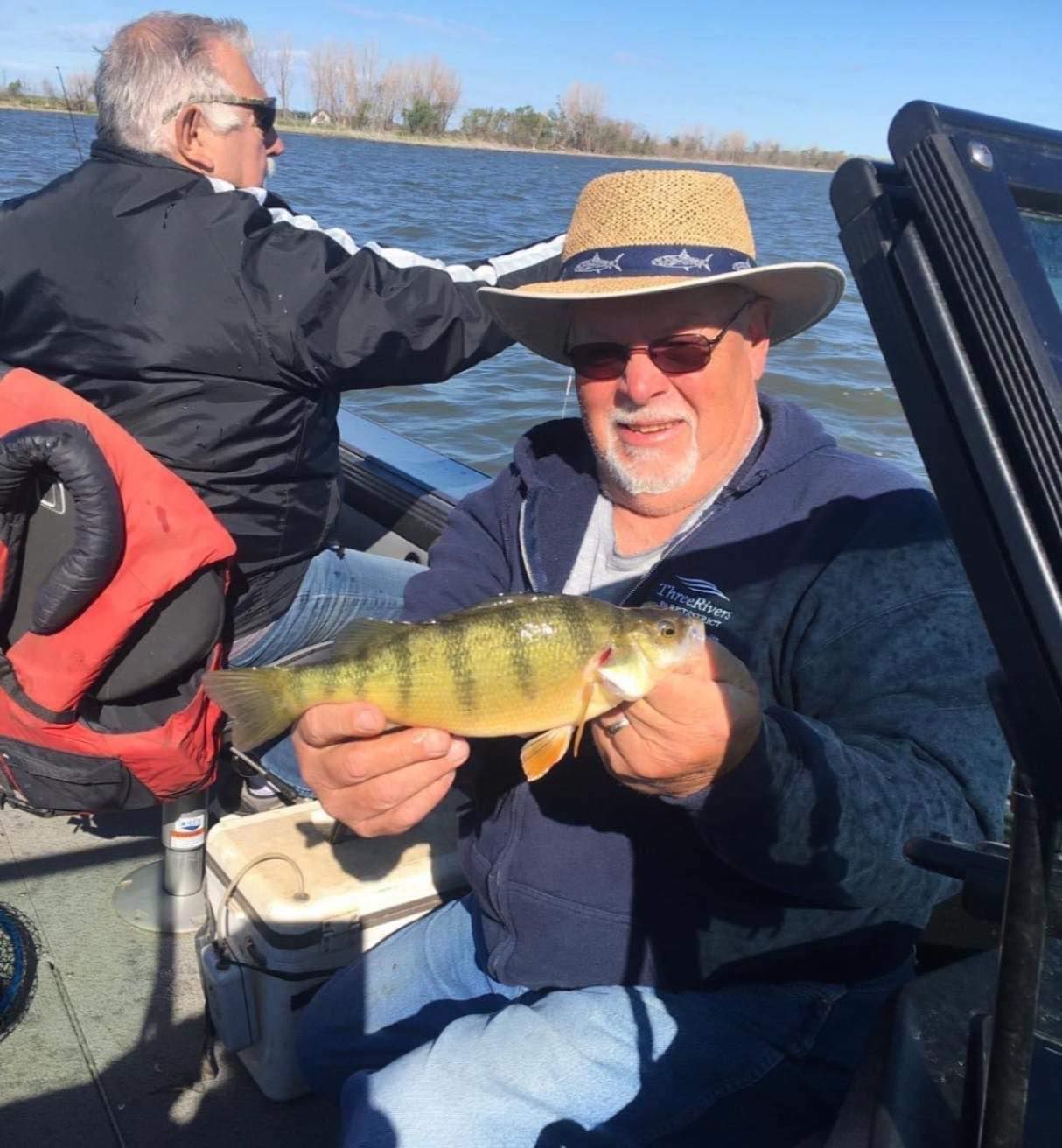 Man on boat holds up a yellow perch fish he caught. Another man sits in the background. Blue water and sky.