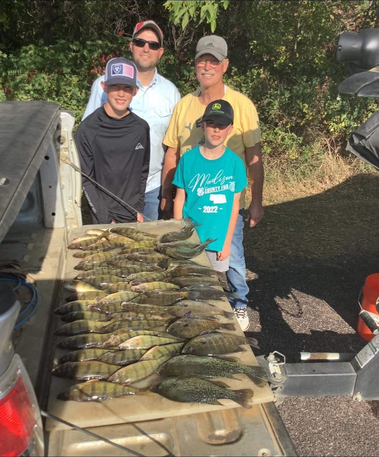 Four people stand by a truck bed filled with fish. Sunlight, green trees in background.