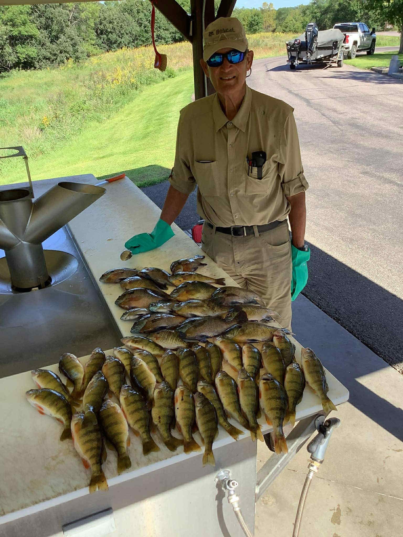 Person in tan clothing, sunglasses, and hat stands with a large pile of yellow perch on a cleaning table near a lake.