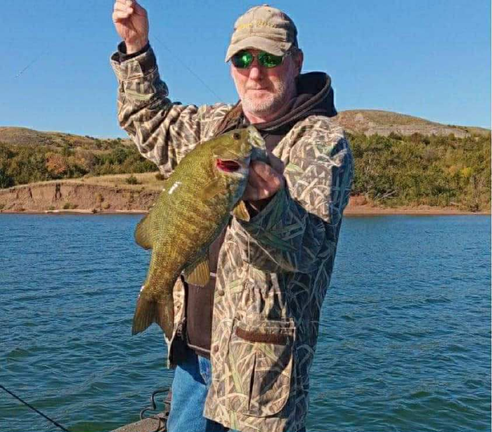 Man in camo holding a large fish, on a boat near a lake, under a blue sky.