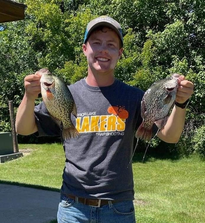 Man holding up two crappie fish, smiling outside. He wears a gray t-shirt and hat. Green grass and trees are in the background.
