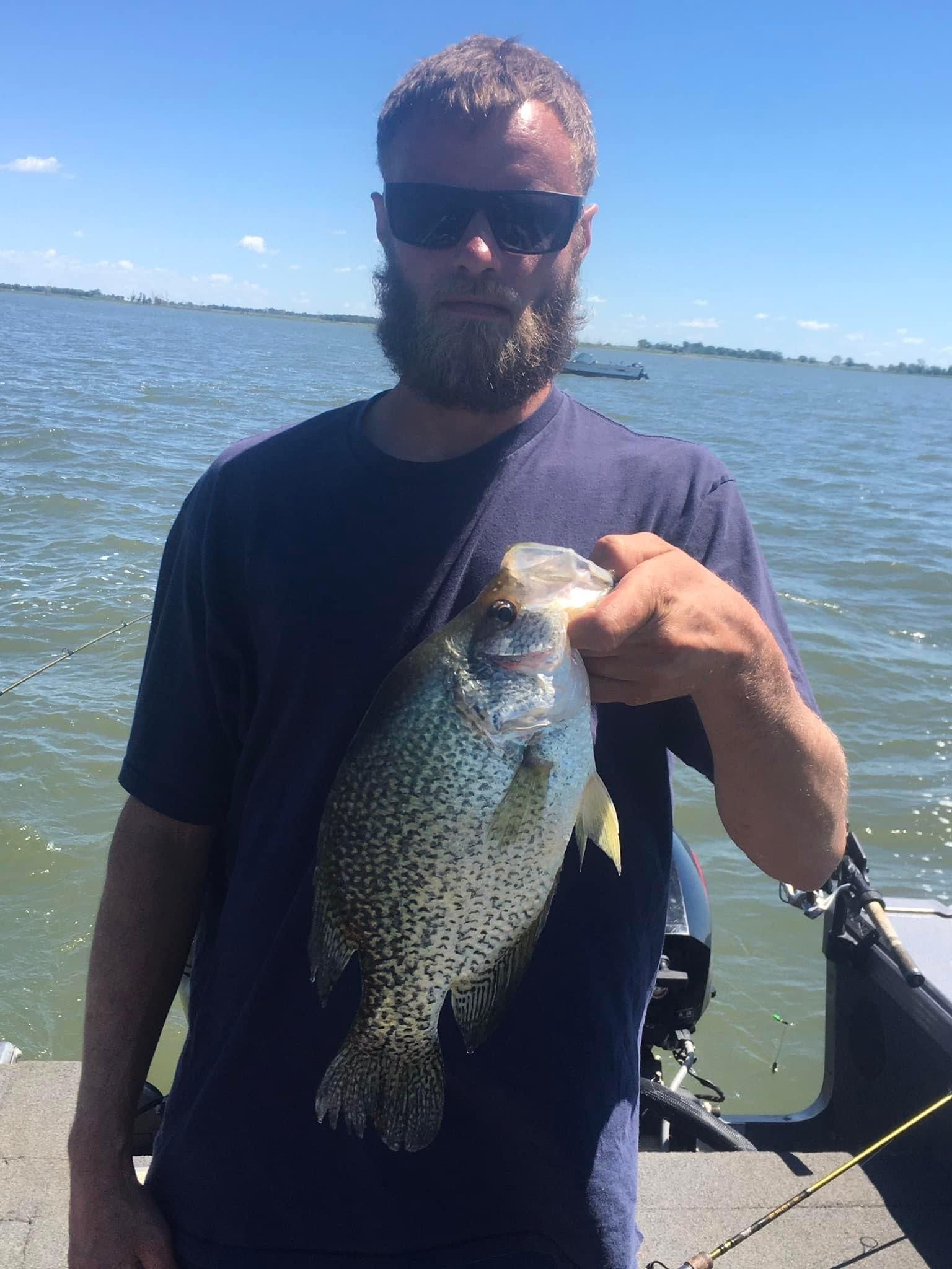 Man on a boat holding a large speckled fish. Water and blue sky in background.