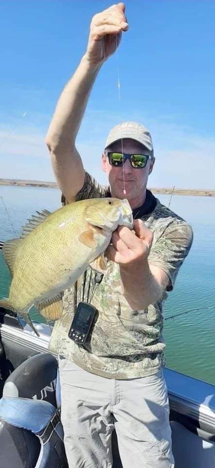 Man in sunglasses holds up a fish on a boat, fishing line dangling. Clear sky and water.