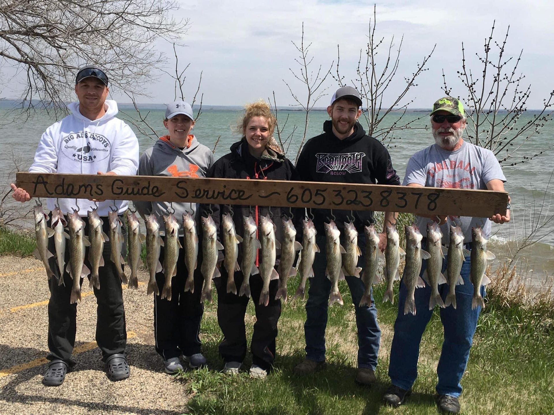 Group of people holding a wooden sign and a string of caught fish near a body of water. Sign has a phone number.